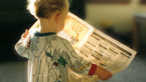 A young child in patterned pajamas holds a newspaper, facing away. Sunlight creates a warm atmosphere in a soft, indoor setting.